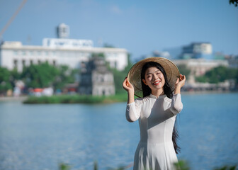 A young Vietnamese woman in a white ao dai and a conical hat standing by Hoan Kiem Lake in Hanoi, Vietnam