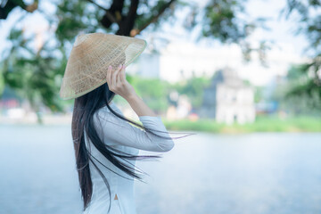 A young Vietnamese woman in a white ao dai and a conical hat standing by Hoan Kiem Lake in Hanoi, Vietnam