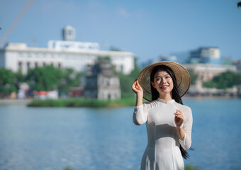 A young Vietnamese woman in a white ao dai and a conical hat standing by Hoan Kiem Lake in Hanoi, Vietnam