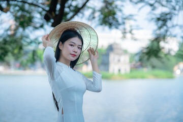 A young Vietnamese woman in a white ao dai and a conical hat standing by Hoan Kiem Lake in Hanoi, Vietnam