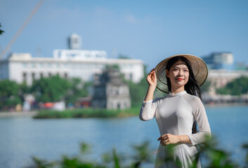 A young Vietnamese woman in a white ao dai and a conical hat standing by Hoan Kiem Lake in Hanoi, Vietnam