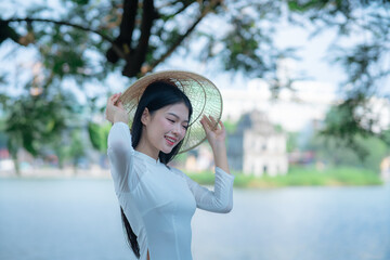 A young Vietnamese woman in a white ao dai and a conical hat standing by Hoan Kiem Lake in Hanoi, Vietnam