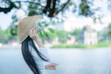 A young Vietnamese woman in a white ao dai and a conical hat standing by Hoan Kiem Lake in Hanoi, Vietnam