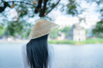 A young Vietnamese woman in a white ao dai and a conical hat standing by Hoan Kiem Lake in Hanoi, Vietnam