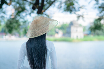 A young Vietnamese woman in a white ao dai and a conical hat standing by Hoan Kiem Lake in Hanoi, Vietnam