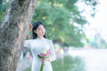 A young Vietnamese woman in a white ao dai standing by Hoan Kiem Lake, holding a bouquet of pink lotus flowers