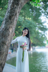 A young Vietnamese woman in a white ao dai standing by Hoan Kiem Lake, holding a bouquet of pink lotus flowers