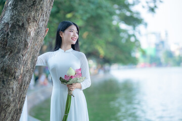 A young Vietnamese woman in a white ao dai standing by Hoan Kiem Lake, holding a bouquet of pink lotus flowers