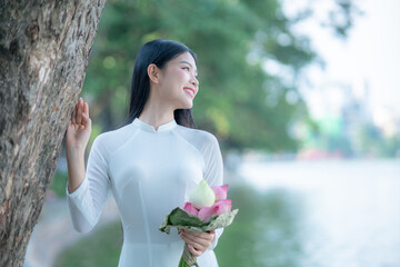 A young Vietnamese woman in a white ao dai standing by Hoan Kiem Lake, holding a bouquet of pink lotus flowers