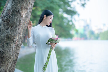 A young Vietnamese woman in a white ao dai standing by Hoan Kiem Lake, holding a bouquet of pink lotus flowers