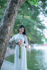 A young Vietnamese woman in a white ao dai standing by Hoan Kiem Lake, holding a bouquet of pink lotus flowers