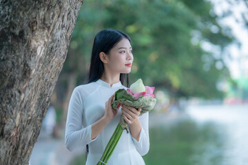 A young Vietnamese woman in a white ao dai standing by Hoan Kiem Lake, holding a bouquet of pink lotus flowers