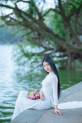 A young Vietnamese woman in a white ao dai standing by Hoan Kiem Lake, holding a bouquet of pink lotus flowers