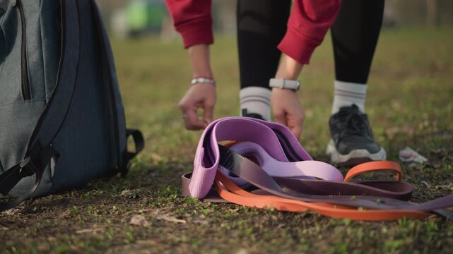 Asian Woman Kneeling In Park Tying Shoelaces Beside Backpack And Stacked Leashes, CloseUp Hands Securing Knot, Athletic Watch Visible, Preparing For Jog Or Dog Walk In Crisp Outdoor Air
