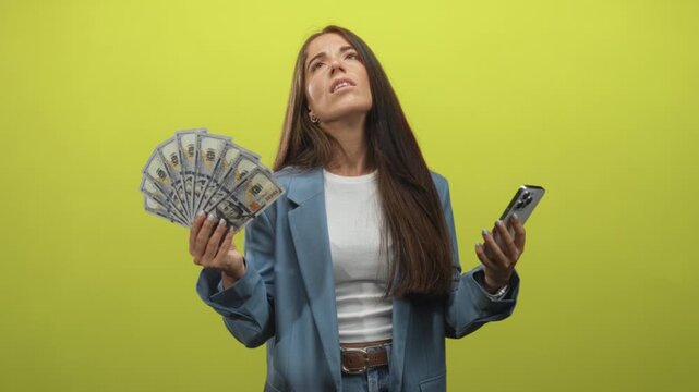 Woman holding a fan of dollars and a smartphone, hands visible, scowling in a studio with bright green backdrop; frustration financial stress.