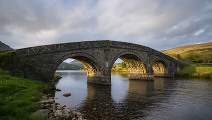 Fototapeta premium Ancient stone bridge covered in moss with soft morning fog, historic weathered architecture surrounded by nature creating a peaceful timeless rural landscape scene