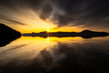 雄大な夜明けの湖。日の出の光と雲の空が湖面に反射するドラマチックな自然の場面。