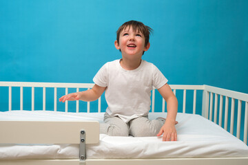 Happy child sitting in white bed on bright blue background. Cute little kid in bed. Joyful boy with smile looking up, for advertisement. Child aged 4 years (four years old)