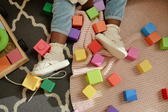 Black toddler sitting on patterned rug surrounded by colorful wooden blocks, wearing socks and shoes, playing with toys, legs and feet visible, engaging in playtime activity