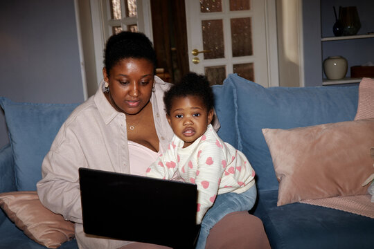 Black woman sitting on sofa using laptop with Black toddler sitting beside her looking toward camera, both engaging with device in home setting, showing family interaction