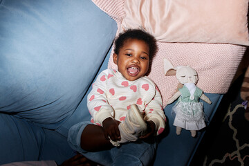 Black toddler girl lying on blue couch smiling and laughing, looking up with plush toy rabbit beside her, showing joyful expression and playful body language