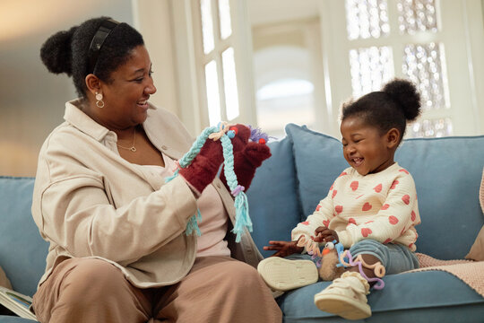 Black woman interacting with Black toddler on sofa, woman holding colorful puppet toys, both smiling and laughing, adult engaging child in playful activity, indoor setting