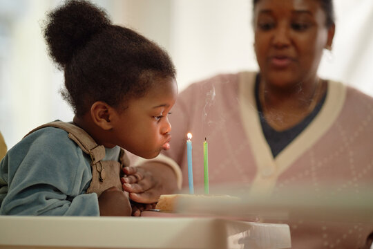 Black child blowing out candles on small cake while Black woman watching closely in background, both sitting at table, celebrating special occasion, focus on childs action
