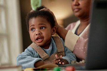 Black woman grooming hair of Black toddler sitting at table, toddler looking away with curious expression, adult partially visible in background, close interaction captured