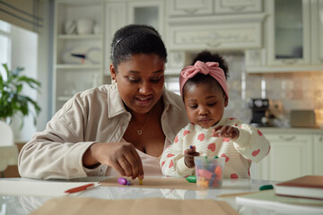 Black woman engaging with Black toddler girl at table, both focused on creative activity with colorful crayons, adult assisting child in developing fine motor skills indoors