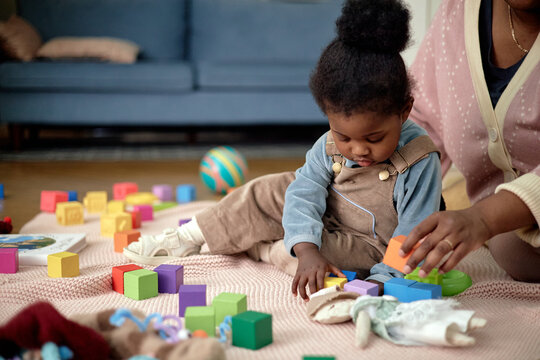 Black woman helping Black toddler playing with colorful building blocks on floor, both sitting on soft blanket, toddler focusing on stacking blocks, adult guiding childs hand during activity