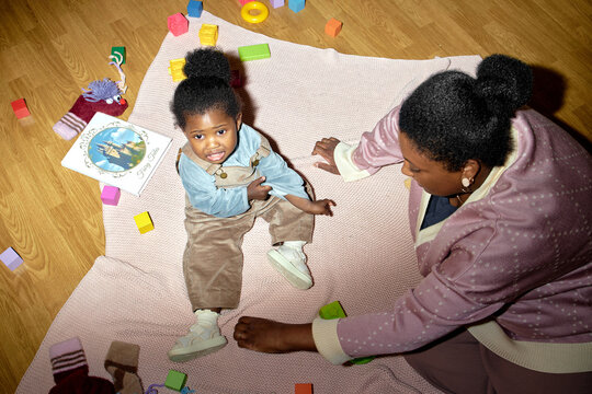 Black woman interacting with Black toddler sitting on blanket surrounded by colorful toys and childrens book, toddler looking up with expressive face, adult engaging child in play