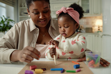 Black woman helping Black toddler drawing with colored pencils and crayons at table, both focused on creative activity