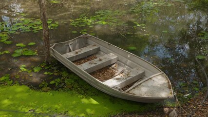 An old fishing tinny boat floats on the murky and mossy water of a lake, tied to the bank with trees shadowing the water and dried leaves filling the boat.