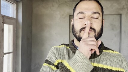Closeup serious caucasian standing in living room showing silence sign holding finger to mouth young man looking in camera demonstrates gesture hush forbids to speak secret information mystery