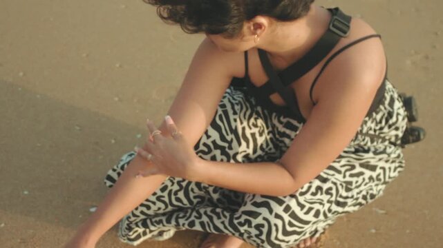 Woman crouching on the sandy beach caressing her arm, enjoying a summer day by the sea, prioritizing skin health and a personal care routine in a windy moment