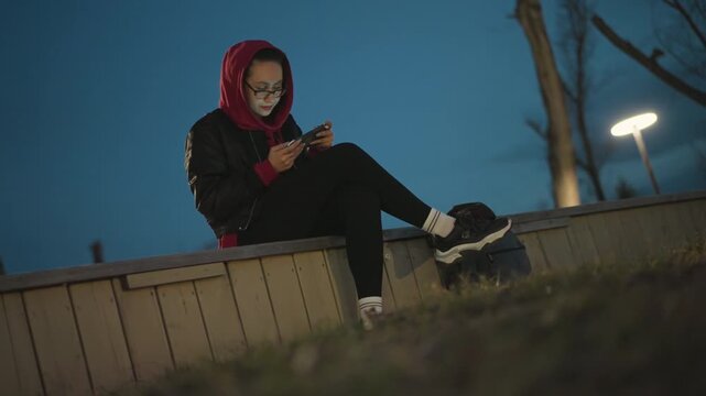 Nighttime Scene Of Woman With Phone In Park, Urban Woman Quietly Reflecting While Writing On Her Phone, Silent Evening In Park As Woman Intently Types On Her Phone Under Soft Lamplight