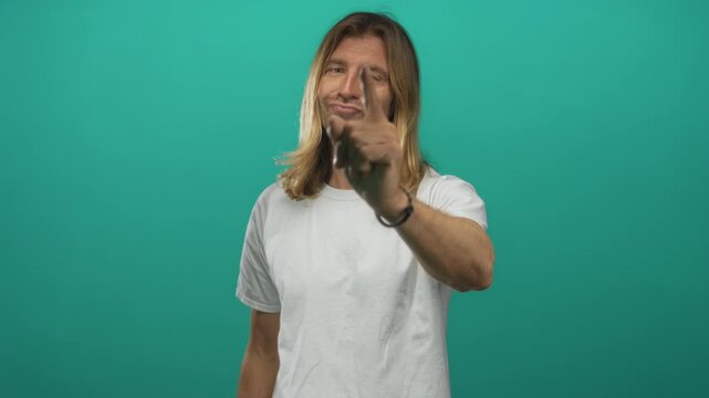 Blond man in white shirt with smug smile boldly points finger toward camera in green studio; confidence.