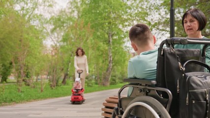 Foreground Wheelchair And Caregiver Observing Child Approaching In Red Toy Car, Anticipation And Gentle Family Dynamic On Park Path, Candid Observation With Green Trees And Soft Light