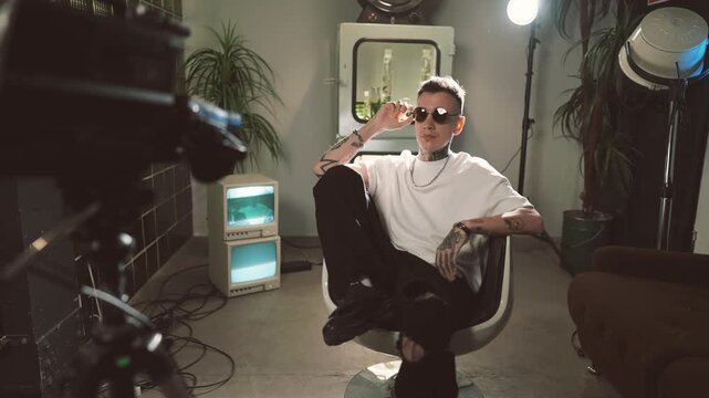 Trendy rap artist posing in studio, shooting music video, portrait indoors. Handsome young male person with tattoos, self-confidence and narcissism, contemporary art interior in studio, modern fashion