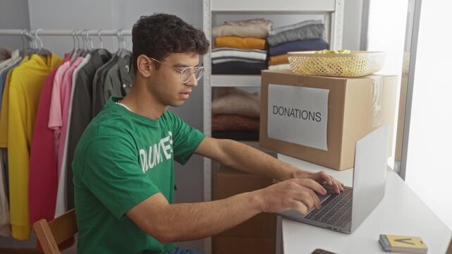 Young man types on laptop beside a cardboard donations box in a building; generosity service compassion.