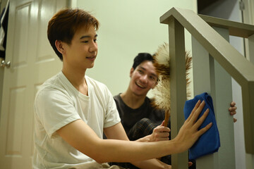 A young Asian gay couple cleans their staircase together, smiling and enjoying the shared moment of doing simple household tasks as a team