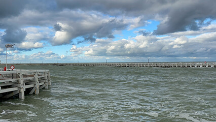 Nieuwpoort, West-Vlaanderen, Belgium, Ocotber 25th, 2025, choppy sea by wooden pier under brooding clouds with distant bridge and lifebuoy on post, turbulent water and textured waves, cool