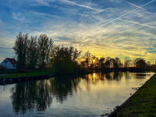 Sunset river reflection with vibrant sky calm water mirroring trees and cottages, contrails streaking across colorful clouds, warm