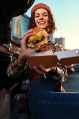 Young woman with burger and bright smile by car in urban outdoor scene, a candid lifestyle portrait capturing authenticity, golden hour glow, mindful living and emotional storytelling in a relaxed