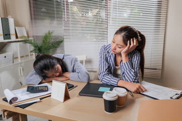 Young women business team work from home office, online learning leads to fatigue, one woman sleeps on desk, other colleague looks weary processing documents, work exhaustion apparent
