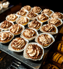 A display of mini chocolate caramel tarts topped with swirled cream and chocolate shavings, arranged in paper cups on a tray, captured in warm lighting for a delicious dessert presentation