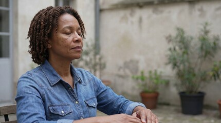Serene middle-aged black woman with closed eyes meditating and practicing mindfulness while sitting outdoors in her backyard patio.