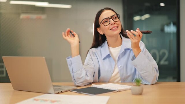 Focused professional records voice message using phone in bright office environment. Displaying contentment and efficiency while taking notes, engaged in modern business communication.