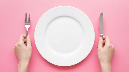Hands holding fork and knife positioned on either side of a blank white plate on a vibrant pink background, suggesting anticipation for a meal