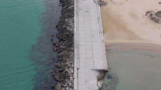Drone footage captures a concrete breakwater surrounded by dark tetrapods in turquoise coastal water, showing calm waves, shoreline details and peaceful marine atmosphere.